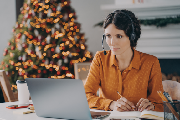 Woman in orange shirt working at laptop with Christmas tree in the background.